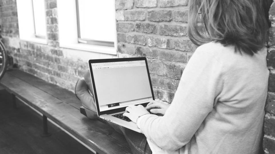 A woman working on a laptop at a long wooden bench beside a brick wall