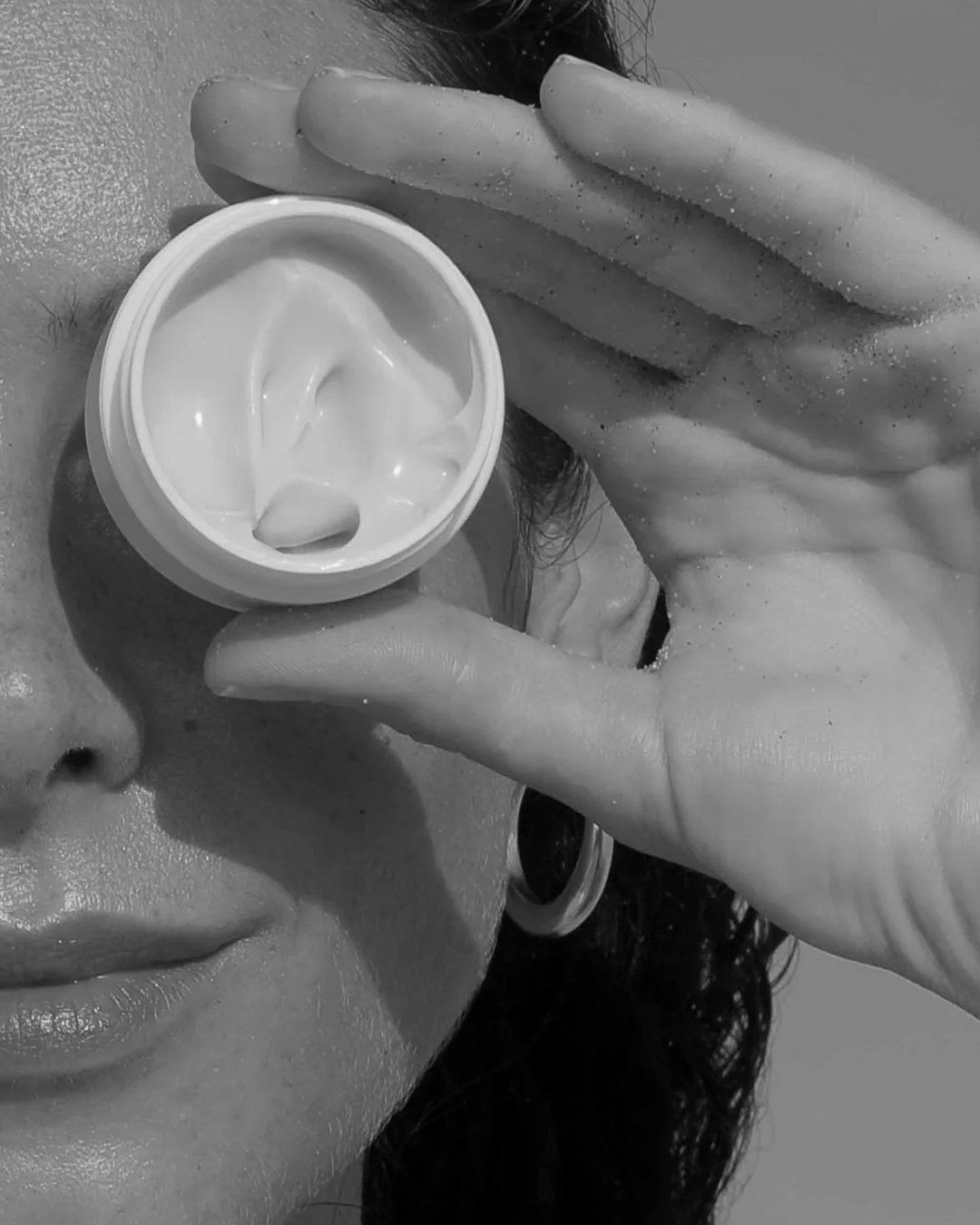 A person holding an open jar of Epi.Logic skincare cream over one eye